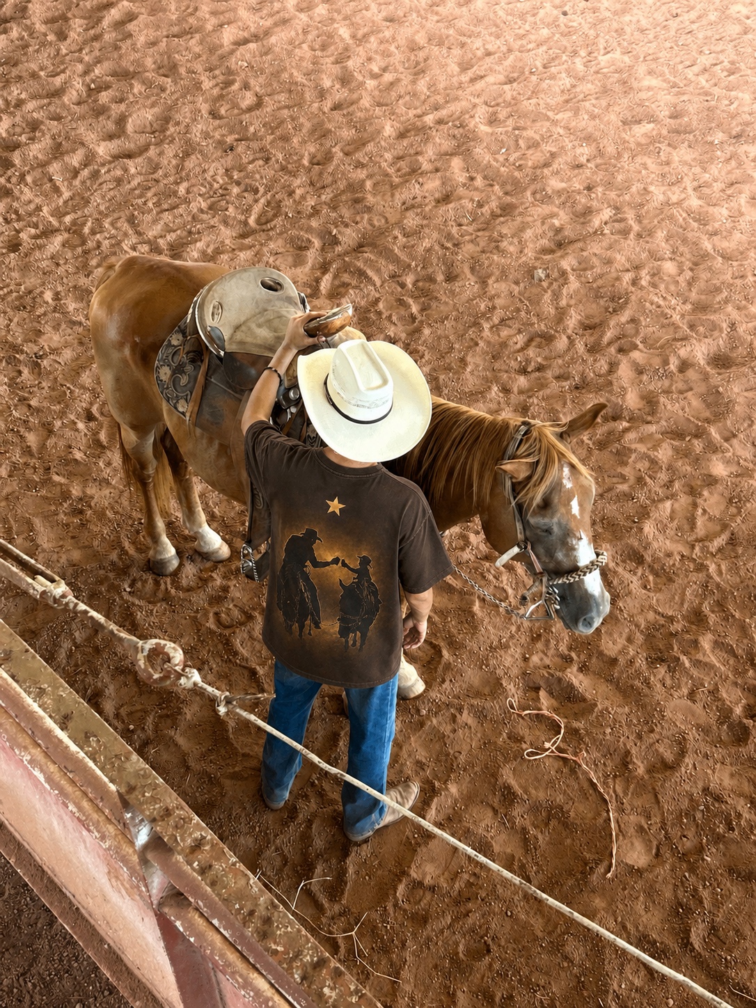 Vaquero con playera ODEL y sombrero junto a un caballo ensillado, vistos desde arriba